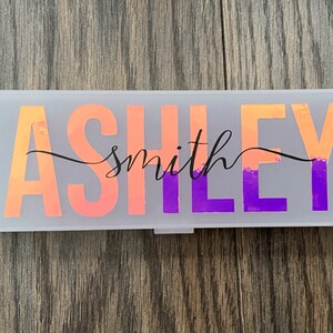 May include: A white plastic pencil case with the name "Ashley smith" written in black cursive script on a pink and purple ombre background.