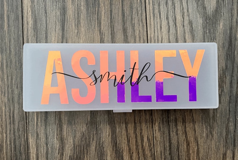 May include: A white plastic pencil case with the name "Ashley smith" written in black cursive script on a pink and purple ombre background.