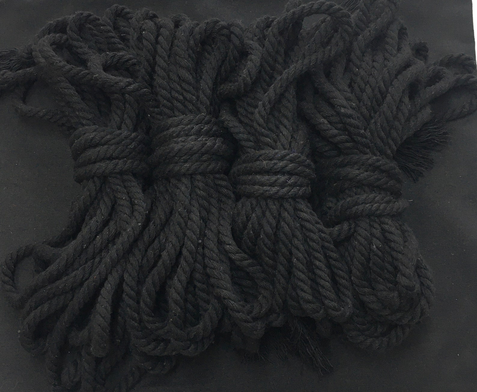 Black cotton shibari ropes coiled on black surface for bondage wear, restraint sex, and handcuff bondage play.