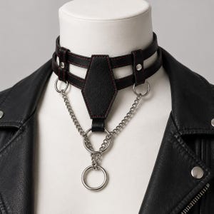 May include: Black leather choker with red stitching and silver-tone chain accents. The choker features a geometric center piece and two parallel straps. A silver chain hangs from the center, connecting to a large ring.