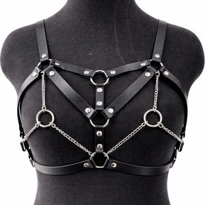 May include: A black harness featuring multiple adjustable straps connected by silver-toned metal rings and studs. The design includes thin vertical and horizontal straps across the chest, accented with delicate silver-toned chains connecting the rings.