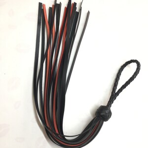 May include: A black and red leather flogger with a braided handle and a knotted base. The flogger features multiple leather strips in black and red, with a braided loop at the end of the handle.