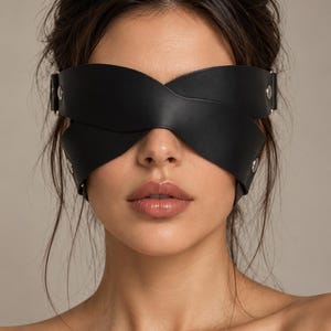 Op de afbeelding: Een zwart lederen blinddoek met een kruislings ontwerp dat de ogen bedekt. Het accessoire is bevestigd met verstelbare zijbanden en zilverkleurige metalen hardware, inclusief ronde klinknagels aan weerszijden van het hoofddeksel.