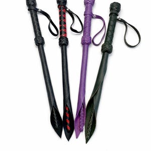 Puede incluir: Cuatro látigos de cuero en negro, rojo, morado y verde. Cada látigo tiene un mango trenzado y una presilla para la muñeca. Los látigos están dispuestos en forma de abanico sobre un fondo blanco.