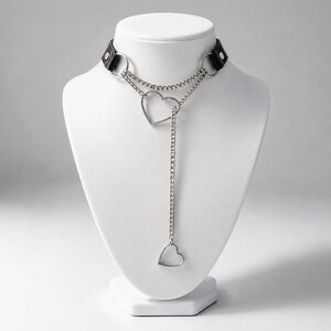 Puede incluir: Un collar gargantilla de cuero negro con cadena de metal plateado y colgantes de corazón. La gargantilla tiene dos anillos plateados y un cierre de hebilla. El collar se exhibe en un busto de maniquí blanco.