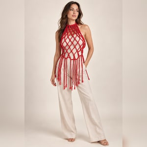 May include: A bright red sleeveless halter top featuring a macrame-style open net design with long fringe detailing at the hem, paired with high-waisted cream-colored wide-leg trousers and simple beige open-toe sandals for a complete outfit.