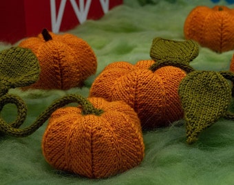 Könnte beinhalten: Fünf gestrickte orangefarbene Kürbisse mit grünen Blättern und Ranken. Die Kürbisse sind auf einer grünen, flauschigen Oberfläche angeordnet.
