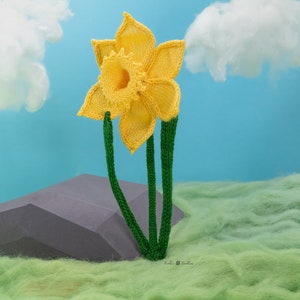 May include: A yellow knitted daffodil flower with green stems against a blue background with white clouds. The flower is sitting on a grey rock and green grass.