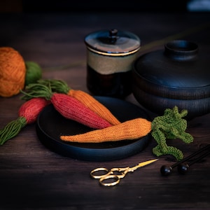 Op de afbeelding: Een set van drie gebreide wortels in oranje, rood en groen, met een zwart bord en een kleine bruine keramische pot met deksel. De wortels zijn op het bord gerangschikt, met de groene wortel bovenop. De pot staat op de achtergrond.