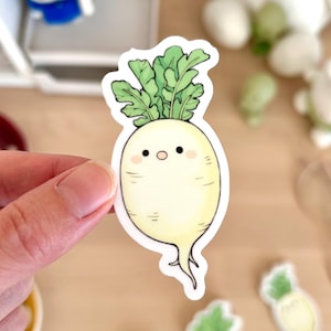 Op de afbeelding: Een witte radijssticker met groene bladeren wordt door een hand vastgehouden. De radijs heeft een schattig gezicht met zwarte ogen en een kleine mond. Andere stickers met vergelijkbare ontwerpen zijn verspreid.