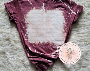 May include: A maroon t-shirt with a bleach-splattered design. The shirt has a round neckline and short sleeves. The text "Vement Living Designs" and "Grow Your Passion" is printed on a white circle with a lightbulb graphic.