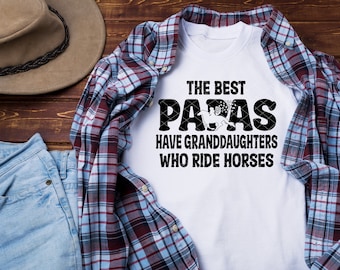 May include: White t-shirt with black text that reads "The Best Papas Have Granddaughters Who Ride Horses". The text is surrounded by a black border. The shirt is laid out on a wooden surface with a blue plaid shirt, a brown hat, and blue jeans.