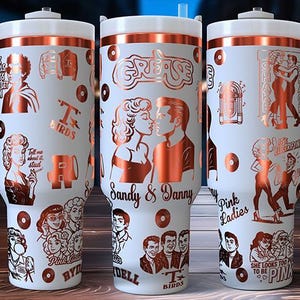 May include: White tumbler with a copper-colored design featuring characters and imagery from the movie Grease. The tumbler has a white handle and lid, and includes the text "Grease", "Sandy & Danny", "Pink Ladies", and "T Birds".
