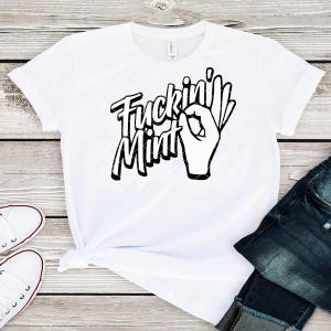 Può includere: T-shirt bianco con la scritta "Fuckin' Mint" in nero, con un font stilizzato. Il design include un gesto della mano. La maglietta è abbinata a jeans blu e scarpe da ginnastica bianche.