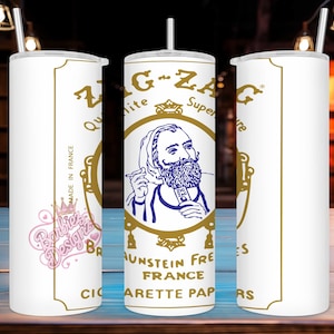 May include: White tumbler with a gold-colored design featuring the text "ZIG-ZAG" and an illustration of a man with a beard. The design includes the words "MADE IN FRANCE" and "CIGARETTE PAPERS". The tumbler has a silver straw and lid.