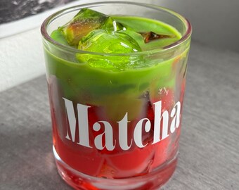 May include: A layered candle in a clear glass, featuring red, green, and clear wax. The word "Matcha" is printed in white on the glass. The top layer has green, gel-like cubes. The candle sits on a gray surface.