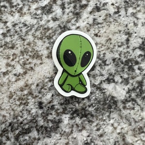 May include: A green alien plush toy with stitched details and black eyes. The alien is sitting with its legs crossed and has a stitched mouth.