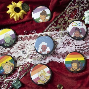 Può includere: Sette spille rotonde con diverse bandiere pride e personaggi dei cartoni animati. Le bandiere includono le bandiere genderqueer, lesbica, bisessuale, transgender, pansexuale e non binaria. I personaggi sono tutti della serie animata She-Ra e le Principesse del Potere.