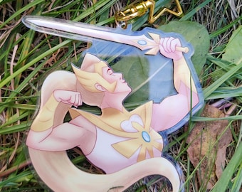 May include: A clear acrylic keychain featuring a cartoon illustration of a blonde woman with a sword. The woman is wearing a gold and white outfit. The keychain has a gold star-shaped clasp and a silver sword.