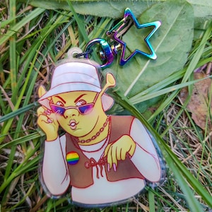 Peut inclure: Un porte-clés en acrylique transparent représentant un personnage de dessin animé avec des lunettes de soleil, un chapeau et un gilet marron. Le porte-clés a un emblème aux couleurs de l'arc-en-ciel et un fermoir en forme d'étoile avec une finition arc-en-ciel.