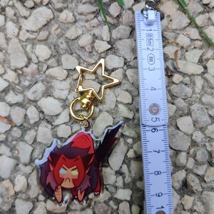 May include: A clear acrylic keychain featuring a cartoon character with red hair, a red and black outfit, and yellow eyes. The character is in a crouching position with a grumpy expression. The keychain is attached to a gold star-shaped charm and a silver ring.