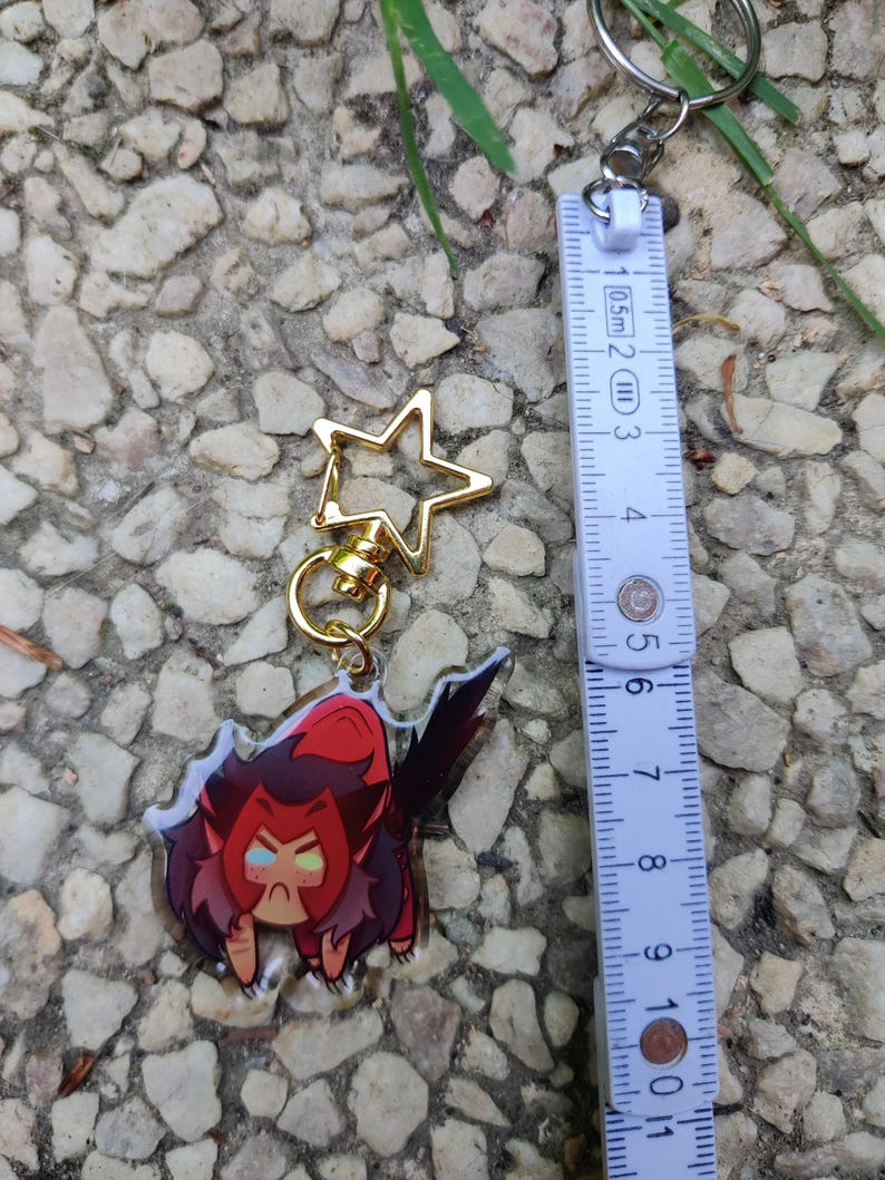 May include: A clear acrylic keychain featuring a cartoon character with red hair, a red and black outfit, and yellow eyes. The character is in a crouching position with a grumpy expression. The keychain is attached to a gold star-shaped charm and a silver ring.