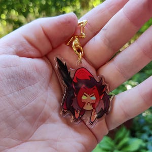 May include: A clear acrylic keychain featuring a cartoon character with red hair, a red and black outfit, and angry facial expression. The character is holding a sword.