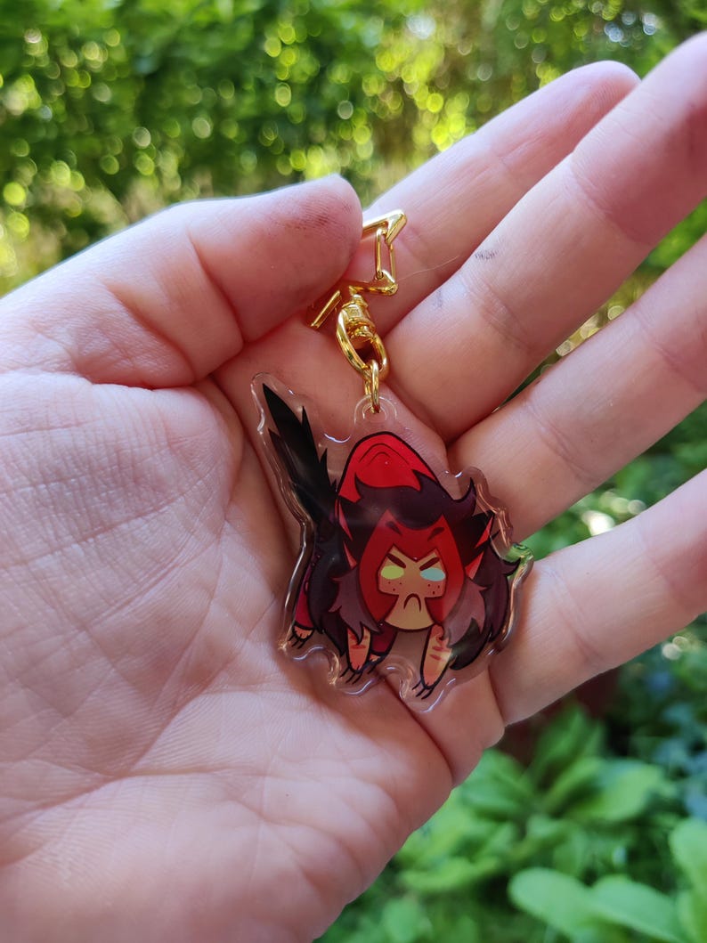 May include: A clear acrylic keychain featuring a cartoon character with red hair, a red and black outfit, and angry facial expression. The character is holding a sword.