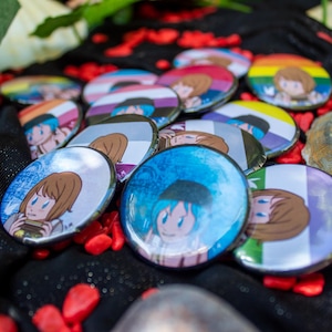 May include: A selection of circular badges featuring anime-style character art. The badges display diverse designs, including rainbow and flag-themed backgrounds. They are arranged on a black fabric with red decorative stones.