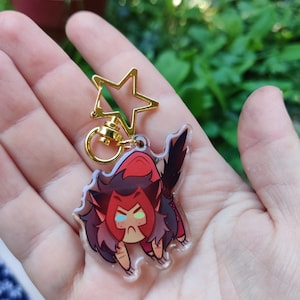 May include: A clear acrylic keychain featuring a cartoon character with red hair, a red and black outfit, and green eyes. The character is in a crouching position with its claws extended. The keychain is attached to a gold star-shaped charm.