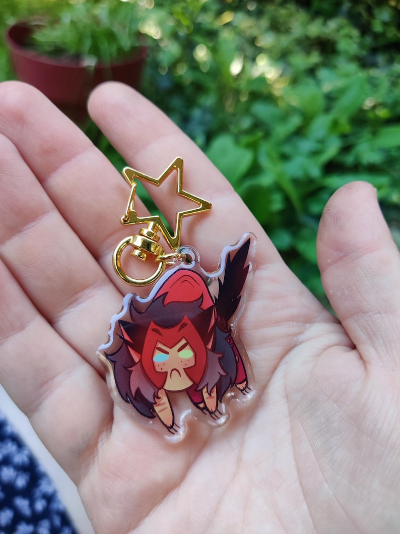 May include: A clear acrylic keychain featuring a cartoon character with red hair, a red and black outfit, and green eyes. The character is in a crouching position with its claws extended. The keychain is attached to a gold star-shaped charm.