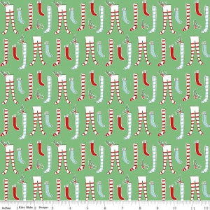 May include: A green fabric with a repeating pattern of red and white striped stockings and blue and white striped stockings. The stockings are arranged in a vertical pattern with a small green sprig of holly between each pair.