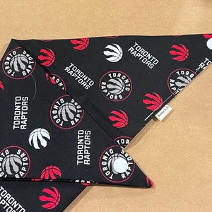 Può includere: Due bandane in tessuto nero con un motivo a logo Toronto Raptors bianco e rosso. Le bandane sono piegate a forma triangolare e hanno un bottone bianco su ogni angolo.