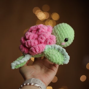 May include: A crocheted pink and green turtle toy with a cute face. The turtle is made with soft yarn and has a fluffy texture.