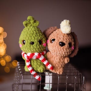 Può includere: Due giocattoli di peluche all'uncinetto, uno verde con una sciarpa a righe rosse e bianche e l'altro marrone, seduti su una griglia metallica.