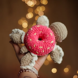 Peut inclure: Un jouet en forme de beignet crocheté rose et blanc avec des bras et des jambes en crochet marron et blanc.