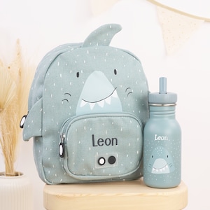 May include: A light blue shark-shaped backpack with a front pocket and a matching light blue water bottle with a straw lid. The backpack and bottle are personalised with the name "Leon".