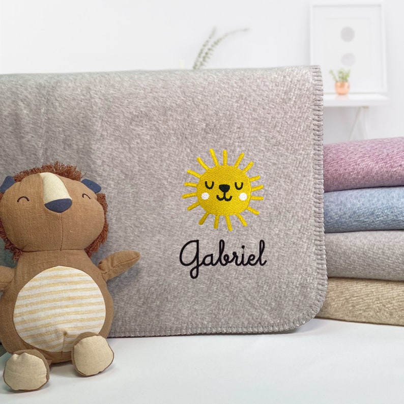 May include: A gray fleece baby blanket with a yellow sun with a face embroidered on it. The name "Gabriel" is embroidered below the sun.