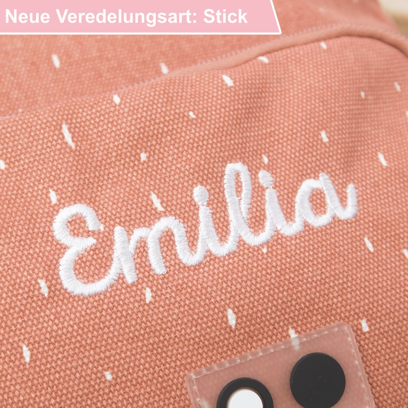 May include: A pink canvas bag with white polka dots and the name "Emilia" embroidered in white. The bag has a clear plastic pocket with two black buttons.