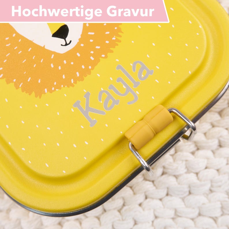 Op de afbeelding: Gele lunchbox met een leeuwenillustratie en de naam "Kayla" gegraveerd op de voorkant. De lunchbox heeft een sluiting en een gele rubberen handgreep.