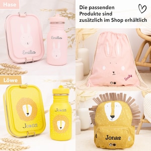 Op de afbeelding: Twee sets gepersonaliseerde lunchboxen, waterflessen en rugzakken. De eerste set is roze met een konijnenontwerp en de naam "Emilia". De tweede set is geel met een leeuwenontwerp en de naam "Jonas".