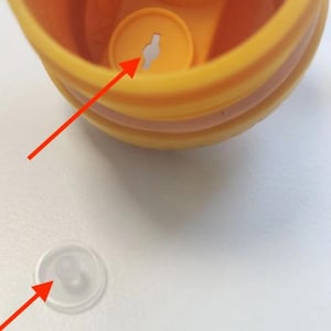May include: A yellow plastic bottle cap with a circular opening featuring a central slot, accompanied by a separate, small, translucent silicone valve plug with a rounded protrusion, both indicated by red arrows for assembly reference.