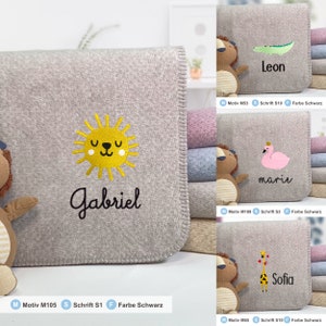 May include: A gray fleece blanket with a yellow sun and the name "Gabriel" embroidered in black. The blanket is folded and stacked with other blankets in different colors.