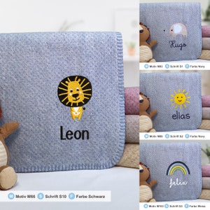 May include: A blue baby blanket with a lion, sun, rainbow, and elephant embroidered on it. The blankets are personalized with the names Leon, Elias, Felix, and Hugo. The text "Motiv M66 Schrift S10 Farbe Schwarz" is at the bottom of the image.