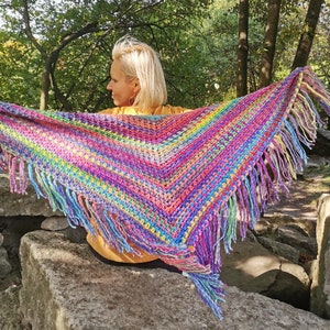 May include: A colorful crocheted shawl with fringe, featuring a rainbow of colors, worn by a person outdoors.