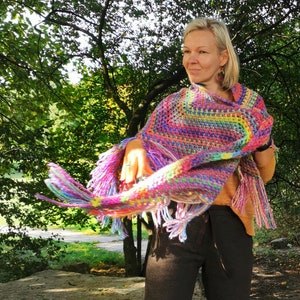 May include: A woman wearing a colorful crocheted poncho with fringe. The poncho is made with a variety of colors, including pink, purple, blue, green, and yellow. The woman is standing in a park with trees in the background.