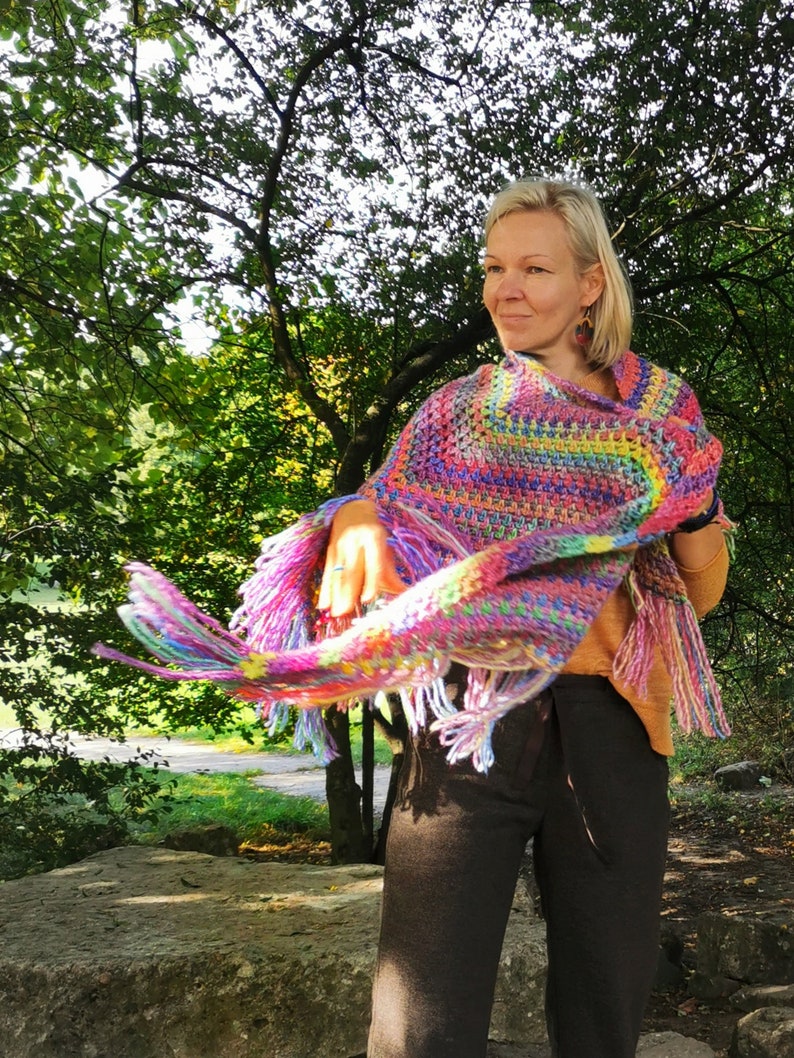 May include: A woman wearing a colorful crocheted poncho with fringe. The poncho is made with a variety of colors, including pink, purple, blue, green, and yellow. The woman is standing in a park with trees in the background.