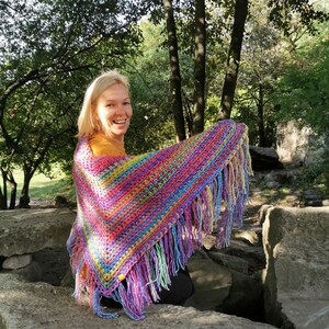 May include: A woman wearing a colorful crocheted shawl with fringe. The shawl is made with a variety of bright colors, including pink, purple, blue, green, and yellow. The woman is smiling and looking at the camera.