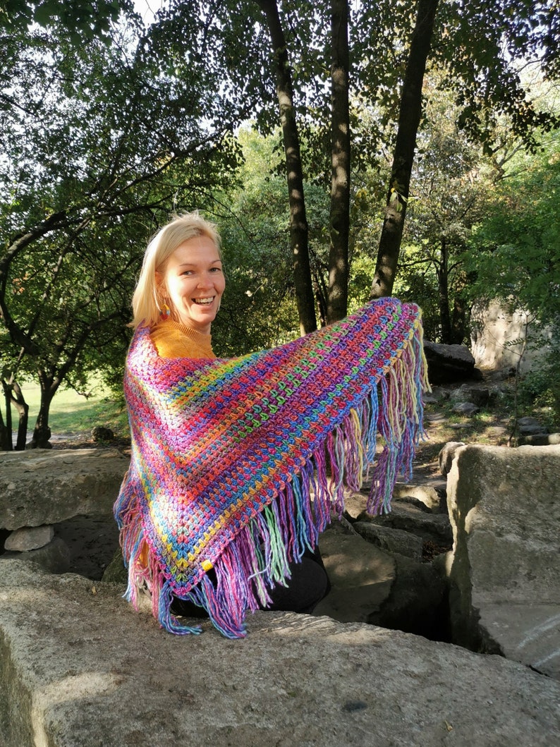 May include: A woman wearing a colorful crocheted shawl with fringe. The shawl is made with a variety of bright colors, including pink, purple, blue, green, and yellow. The woman is smiling and looking at the camera.