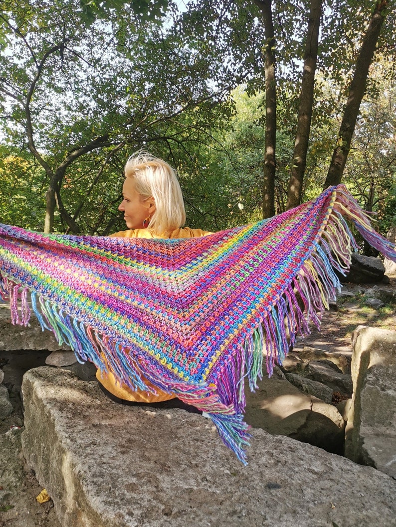 May include: A colorful crocheted shawl with fringe, featuring a rainbow of colors, worn by a person outdoors.